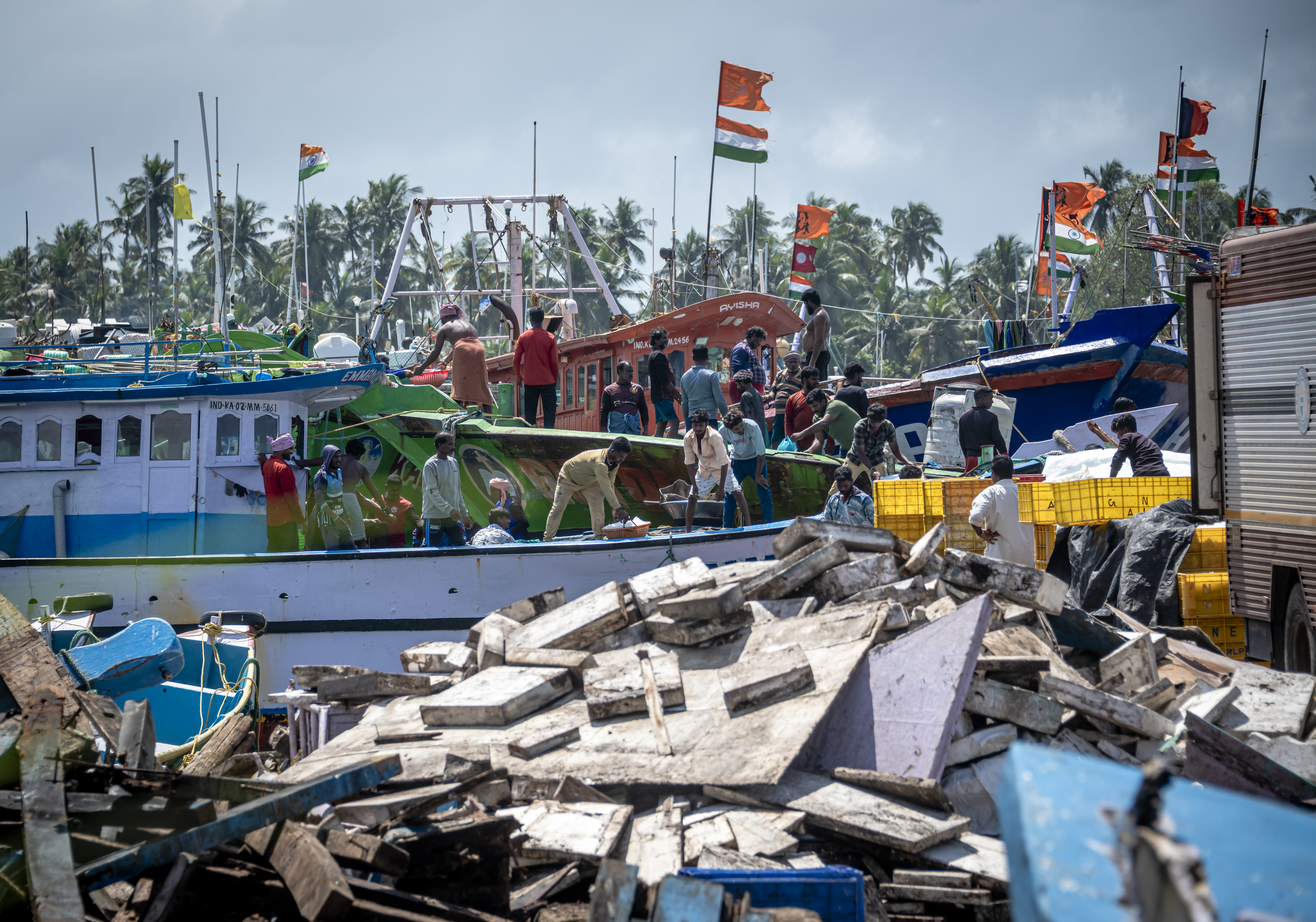 Fishing boats crowded at a harbor with workers loading and unloading various supplies, while colorful flags and palm trees are visible in the background.